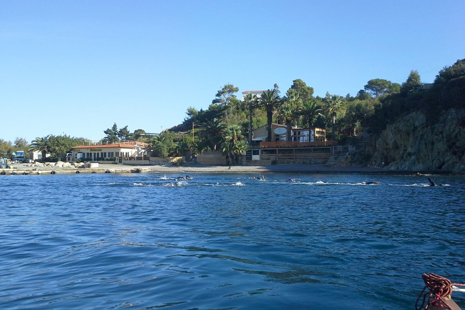 Nuotatori in mare davanti alla spiaggia dell’Enfola con la costa e gli edifici sullo sfondo