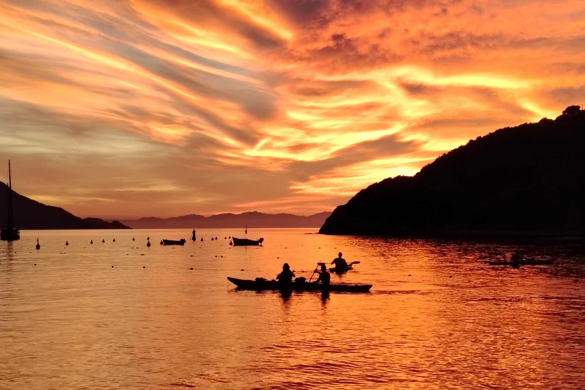 Tramonto sul mare con kayak in controluce, cielo arancione e barche ancorate nella baia