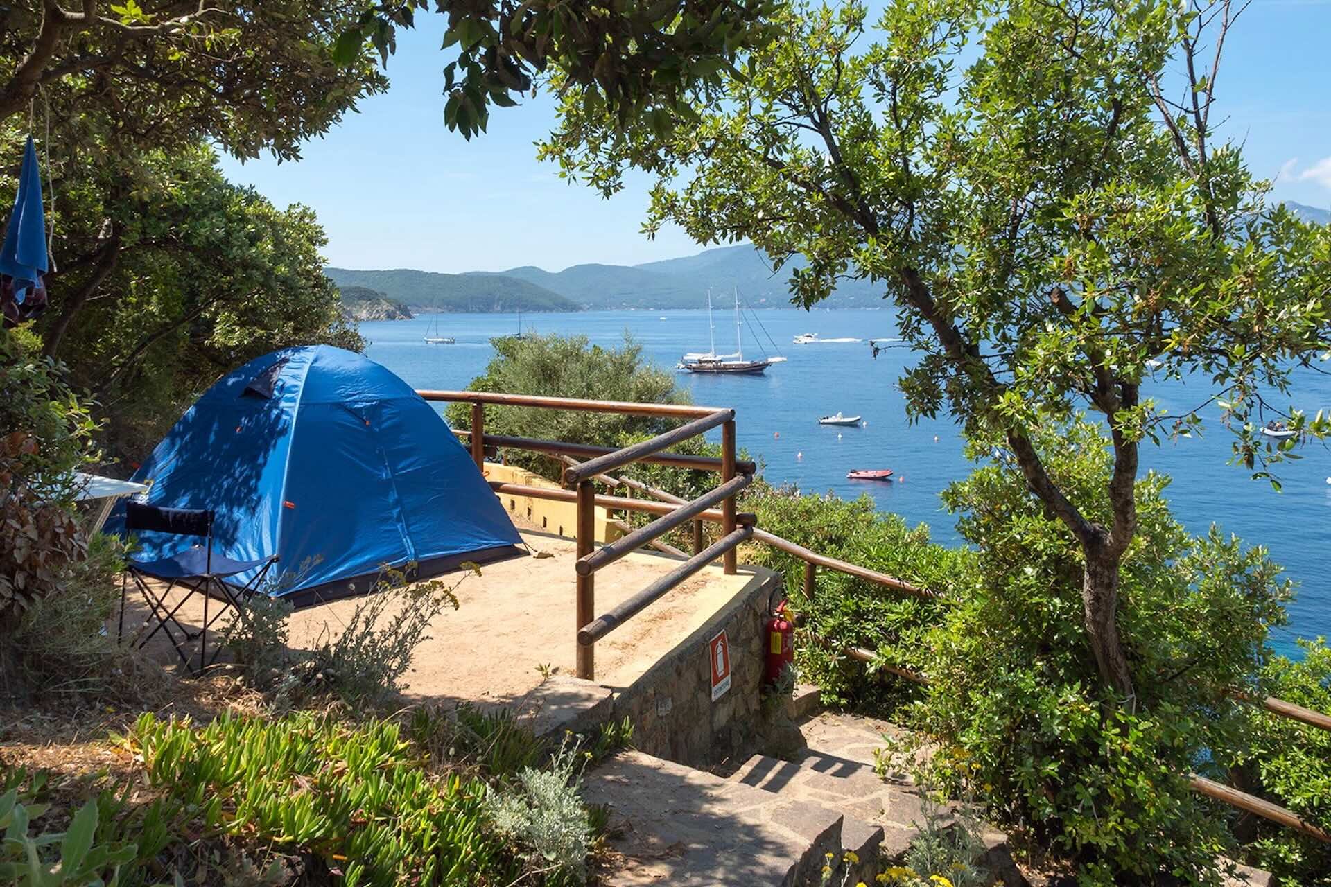 Tenda blu su piazzola terrazzata al Camping Enfola con vista sulla baia e barche ancorate